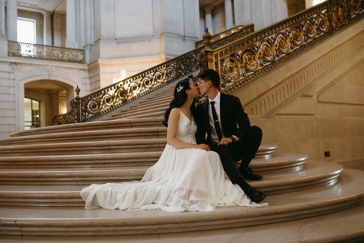 Bride and groom cuddle on steps in San Francisco City Hall on their wedding day.