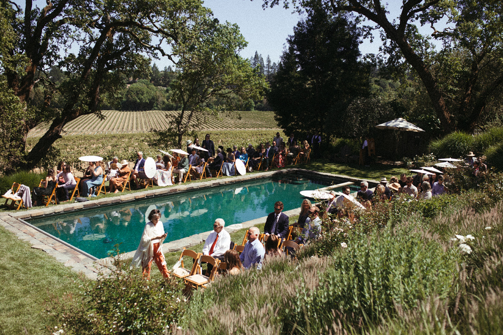 Wedding guests sit around a beautiful blue outdoor pool waiting for the bride and groom.